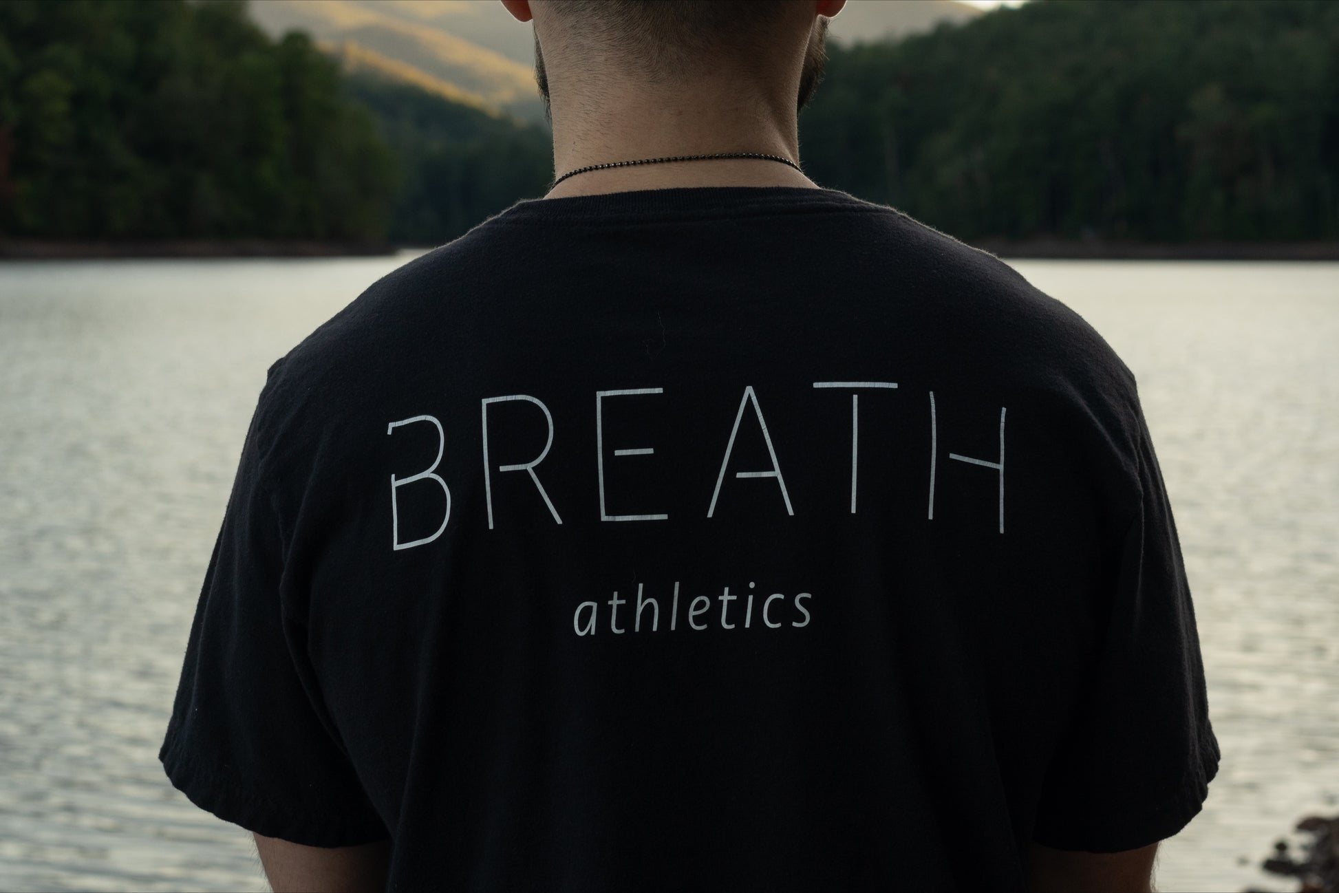 A picture of the upper back of a person person displaying the front of the 100% Organic cotton black graphic t shirt by Breath with the logo on the left chest and the brand name across the back, looking out over a beautiful lake.