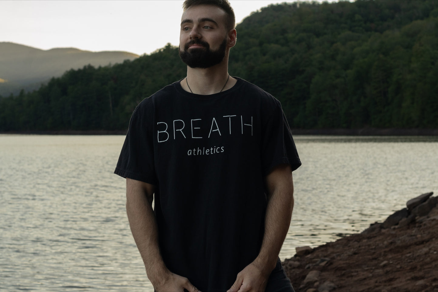 A person Standing in front of a beautiful lake with the black t shirt with the breath logo on the back showing the front of the shirt with the word "Breath".