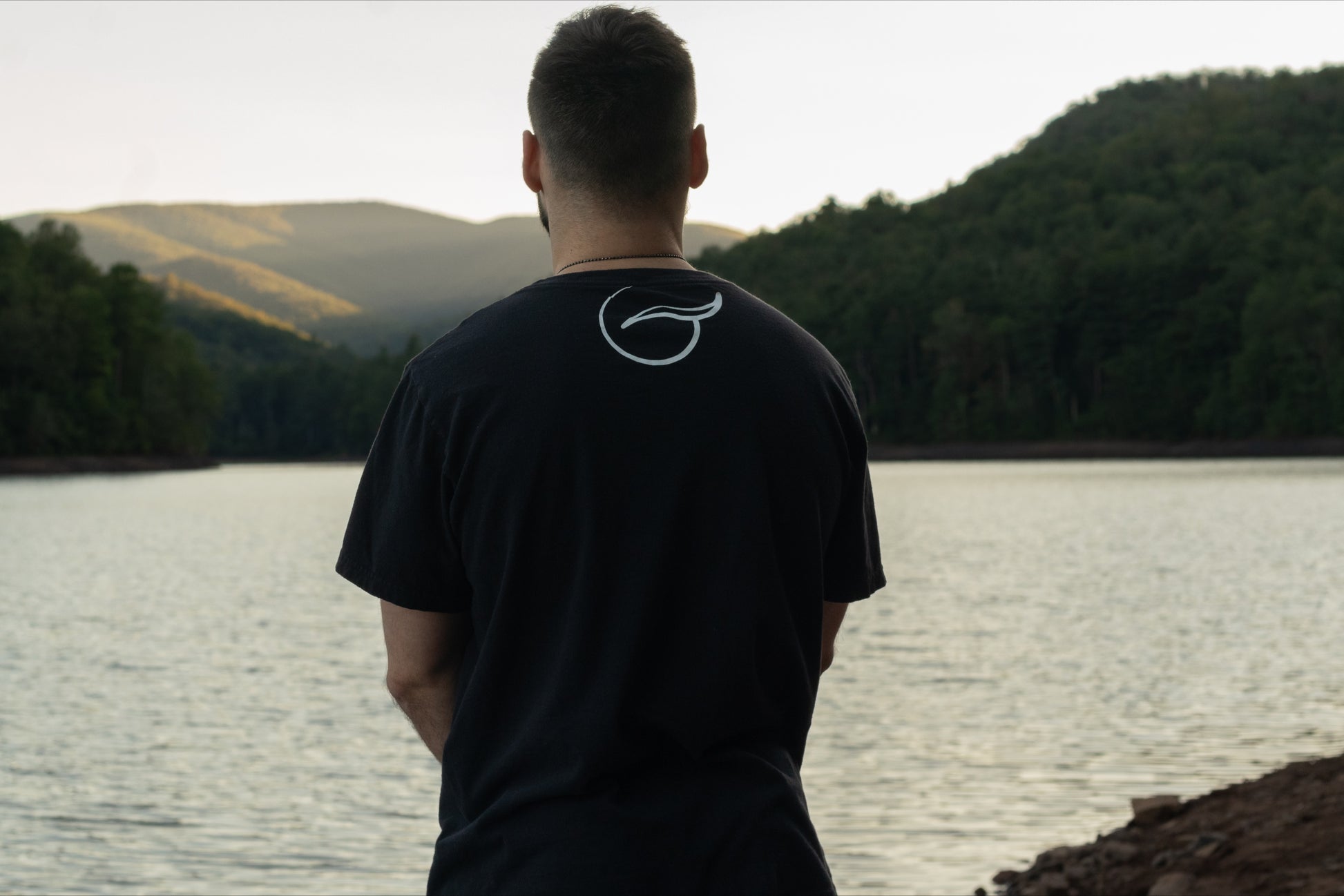 A person facing away looking out over a beautiful lake in the black t-shirt  with the breath logo in the upper center of the back.